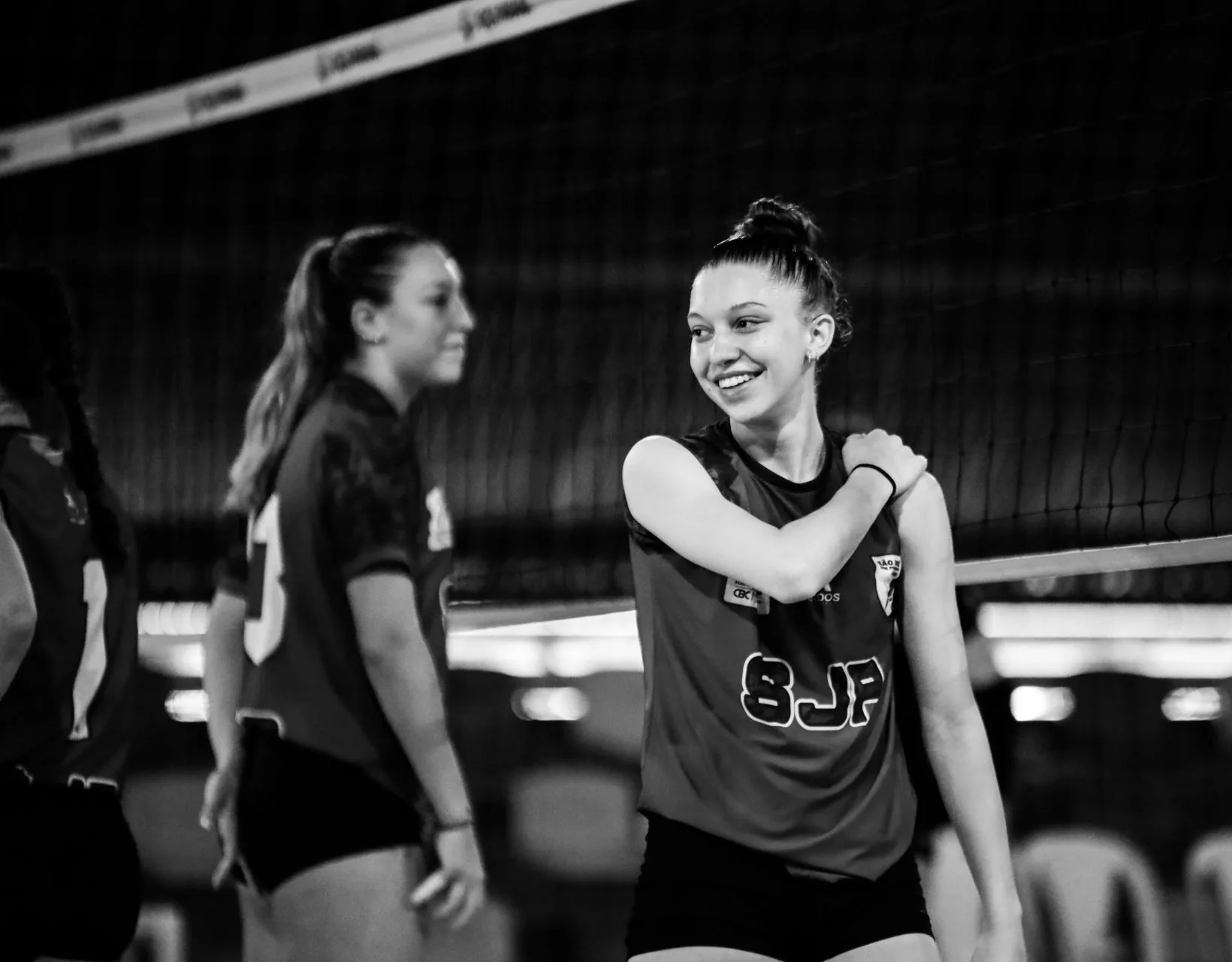 A smiling teenage volleyball player stands at the net, teammates behind her on an indoor court